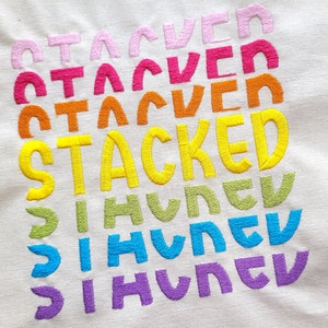 May include: A white fabric with the word "STACKED" embroidered in yellow, with the word "HUNGRY" embroidered in green, blue, and purple below it. The word "STACKED" is repeated three times above it in pink, red, and orange.