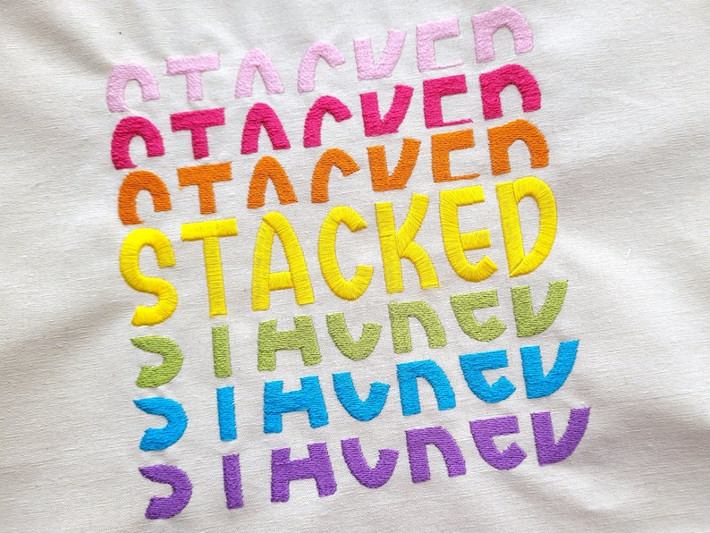 May include: A white fabric with the word "STACKED" embroidered in yellow, with the word "HUNGRY" embroidered in green, blue, and purple below it. The word "STACKED" is repeated three times above it in pink, red, and orange.