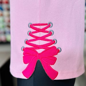 May include: Pink shirt with a pink and grey embroidered corset and bow design.