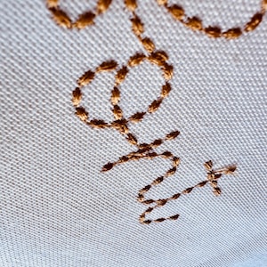 May include: A white fabric surface with a gold embroidered design of a stylized "Love" with a cross at the end.