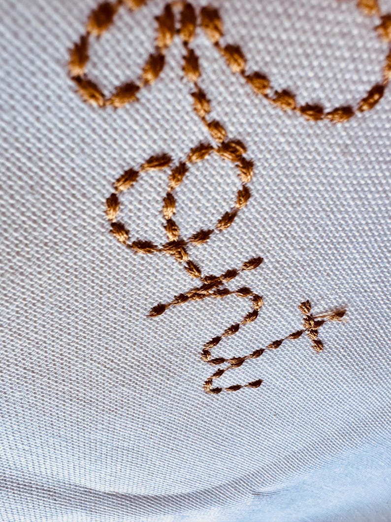 May include: A white fabric surface with a gold embroidered design of a stylized "Love" with a cross at the end.