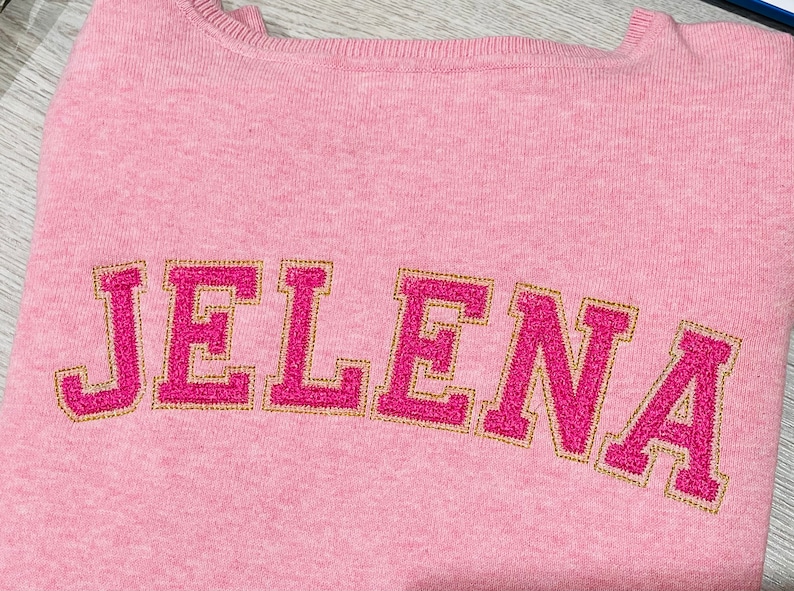 May include: Pink sweater with the name "JELENA" embroidered in pink and gold thread.