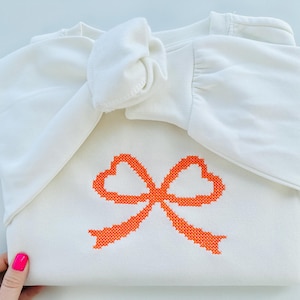 May include: White sweatshirt with a large orange embroidered bow on the front. The sweatshirt is folded in half and the sleeves are tied together in a bow.