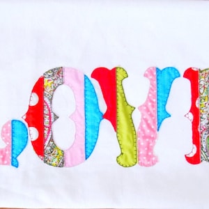 May include: The word "LOVE" is spelled out in large, colorful letters. Each letter is made of different fabric scraps sewn together, creating a patchwork effect. The fabrics include pink, red, blue, green, and white polka dots.