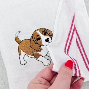 Puede incluir: Toalla de mano blanca con un cachorro beagle bordado. El beagle es marrón, blanco y negro. La toalla tiene rayas rojas en el borde.