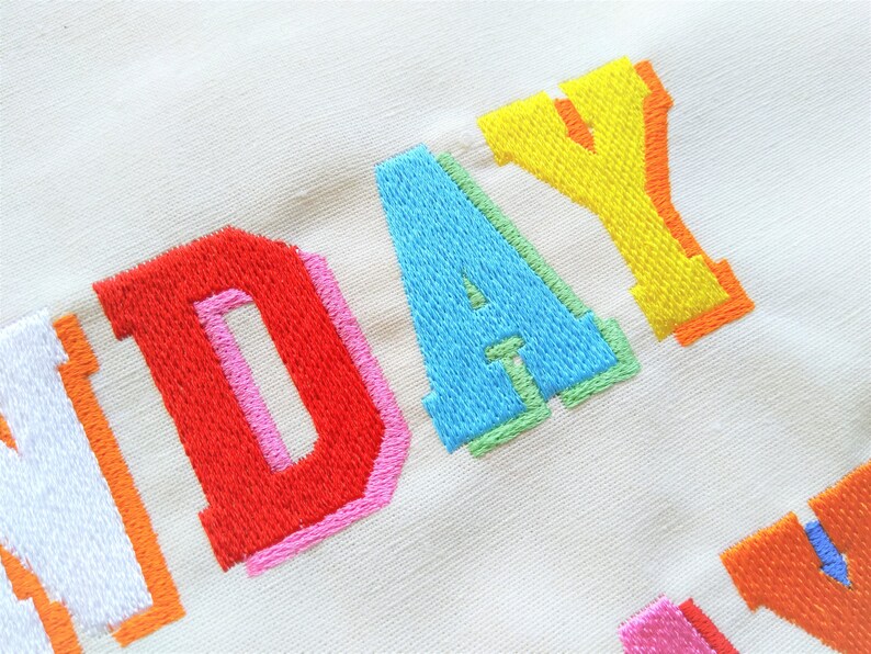 May include: Embroidered word "SUNDAY" in rainbow colors on a white fabric background.