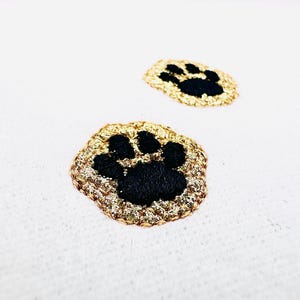 May include: Two embroidered paw print patches featuring black thread for the paw pads and a shimmering gold metallic thread border. The circular patches are stitched with a dense texture, showcasing a clear contrast between the dark center and the bright, reflective gold edge.
