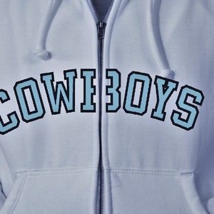 Pode incluir: Um moletom azul claro com zíper e a palavra "COWBOYS" impressa em uma fonte azul e preta.