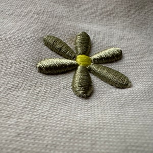 Könnte beinhalten: Eine silberne gestickte Blume mit gelber Mitte auf einem weißen Stoffhintergrund.
