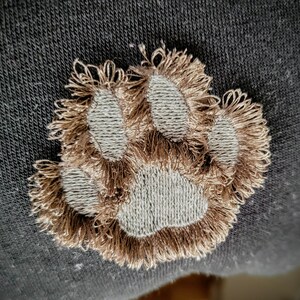 May include: A brown embroidered paw print on a dark gray fabric background.
