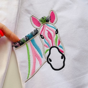 May include: A white sweatshirt with a colorful embroidered zebra design. The zebra has pink, green, and blue stripes.