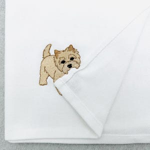 May include: White folded towel featuring an embroidered tan Cairn Terrier dog. The dog is facing forward with its tail raised. The towel has a textured weave.