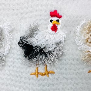 Puede incluir: Pollos bordados en hilo blanco, negro y beige con crestas rojas y patas amarillas. Los pollos están sobre un fondo de punto blanco. La imagen es un primer plano del bordado.