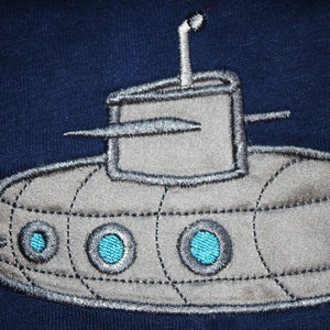 May include: Embroidered patch of a gray submarine with a black propeller and a periscope. The submarine has three portholes with teal blue centers. The patch is sewn onto a navy blue fabric background.