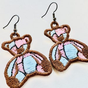 May include: Embroidered teddy bear earrings. The earrings have a brown outline with pink and light blue sections. The ear hooks are dark metal. These earrings are a fashion accessory, designed to be worn.