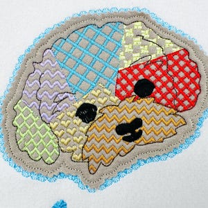 May include: Embroidered patch in the shape of a brain, featuring various patterned sections in blue, green, purple, yellow, and red. The patch has a gray border with a blue scalloped edge. Black details form the eyes and mouth.