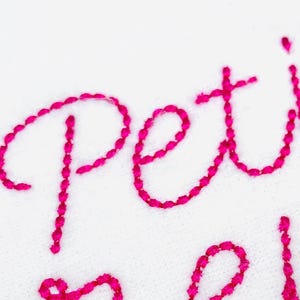 May include: Close-up of white fabric with embroidered text in bright pink. The word "petit" is visible, along with a portion of another word. A small floral design with red and pink petals and green leaves is in the corner.