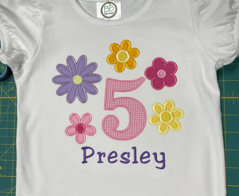 May include: White t-shirt with pink, yellow, and purple embroidered flowers, a pink gingham number "5", and the name "Presley" in purple embroidery.