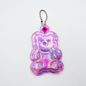 May include: A pink, iridescent gummy bear-shaped keychain. The keychain has a silver chain and a pink eyelet. The gummy bear design is outlined with pink stitching and has a shiny, holographic finish.