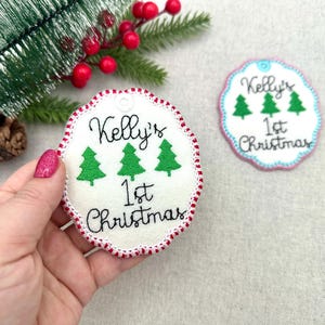 May include: Embroidered Christmas ornament with the text "Kelly's 1st Christmas" and three green Christmas trees. The ornament has a white background with a red and white stitched border. A second ornament is visible.