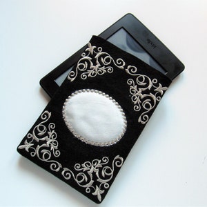 May include: Black felt e-reader case with silver embroidery of a floral design and a white oval insert. The case is shown with a Kindle e-reader inside.