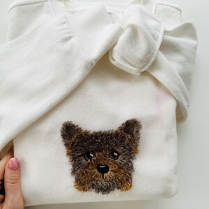 Puede incluir: Sudadera blanca con un rostro de perro bordado en marr&oacute;n y negro.