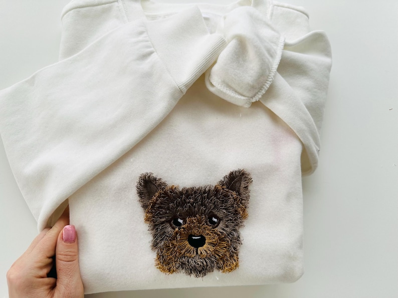 Puede incluir: Sudadera blanca con un rostro de perro bordado en marr&oacute;n y negro.
