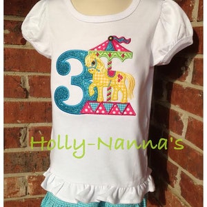 K&ouml;nnte beinhalten: Wei&szlig;es T-Shirt mit einer blauen Zahl 3 und einem bunten Karussellpferd-Design. Das Shirt hat einen R&uuml;schen-Saum und wird mit einem blau-wei&szlig; karierten Rock kombiniert. Der Text "Holly-Namma's" ist auf das Shirt gestickt.