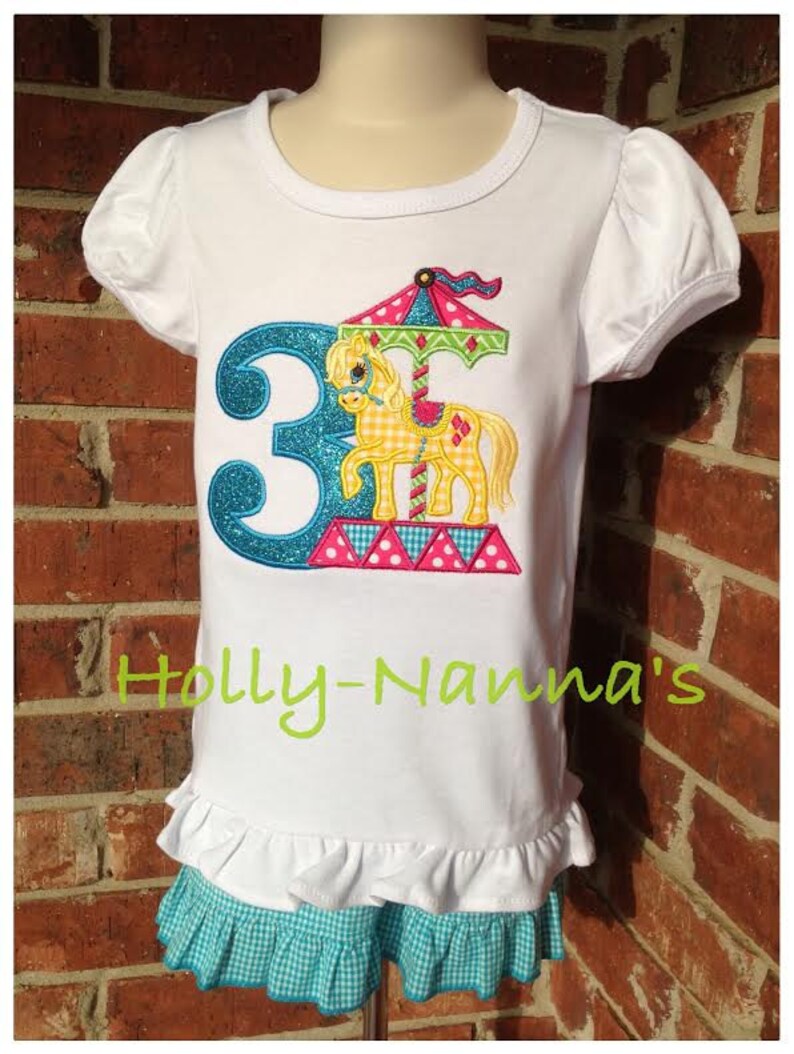K&ouml;nnte beinhalten: Wei&szlig;es T-Shirt mit einer blauen Zahl 3 und einem bunten Karussellpferd-Design. Das Shirt hat einen R&uuml;schen-Saum und wird mit einem blau-wei&szlig; karierten Rock kombiniert. Der Text "Holly-Namma's" ist auf das Shirt gestickt.
