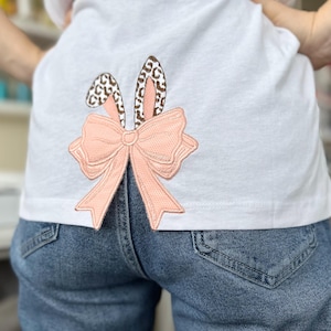 以下が含まれることがあります： 後ろにピンクのリボンと茶色のウサギの耳が刺繍された白いTシャツ。Tシャツは青いジーンズと合わせて着られています。