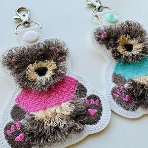 Può includere: Due portachiavi di peluche orsacchiotto ricamati. Uno indossa una camicia rosa e l'altro una camicia turchese. Entrambi hanno la pelliccia marrone e le impronte delle zampe rosa.