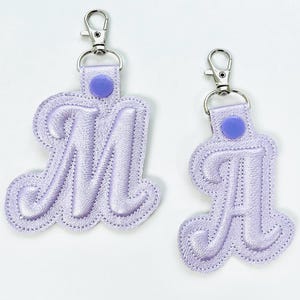 May include: Two lavender keyrings, one with the letter "M" and the other with the letter "A". Each keyring has a silver clasp and a matching lavender button. The letters are embroidered with a decorative script font.