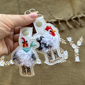 Könnte beinhalten: Zwei weiße Stoffschlüsselanhänger mit gestickten Hühnern, die Stiefel tragen. Die Hühner haben rote Kämme und weiße, flauschige Federn.