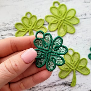 May include: Four green four-leaf clover shapes made of lace. The clovers are different sizes and shades of green. One clover is being held in a hand.