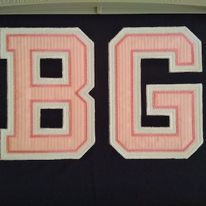 May include: Two large white felt letters, "B" and "G", with pink outlines. The letters are stitched together and have a textured appearance.