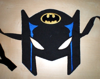 Puede incluir: Máscara de Batman de fieltro negro con un símbolo de murciélago dorado y detalles azules. La máscara tiene agujeros para los ojos y una correa para llevarla.