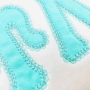 Könnte beinhalten: Nahaufnahme eines weißen Stoffes mit den Buchstaben "RM", die in einem hellen Türkis gestickt sind. Die Buchstaben haben einen gewellten Rand und sind mit einem kontrastierenden weißen Faden genäht, wodurch ein texturierter Effekt entsteht. Das Design ist schlicht und elegant.