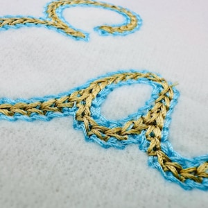Puede incluir: Primer plano de la letra "S" bordada en hilo dorado y azul claro sobre una tela blanca. La costura tiene un aspecto trenzado, con el hilo dorado delineado por el azul claro. La tela tiene una superficie texturizada y ligeramente borrosa.