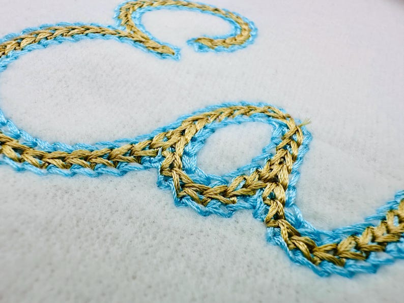 Puede incluir: Primer plano de la letra "S" bordada en hilo dorado y azul claro sobre una tela blanca. La costura tiene un aspecto trenzado, con el hilo dorado delineado por el azul claro. La tela tiene una superficie texturizada y ligeramente borrosa.
