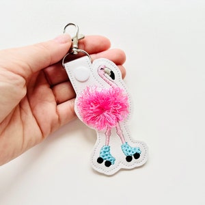 Könnte beinhalten: Weißer Leder-Schlüsselanhänger mit einem rosa Flamingo, der Rollschuhe trägt. Der Flamingo hat einen rosa, flauschigen Tutu und blaue Rollschuhe.