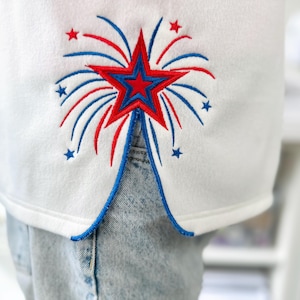 May include: White garment with a red and blue embroidered starburst design. The design features a red star with blue accents and red and blue lines radiating outwards, resembling fireworks. The garment is paired with blue jeans.