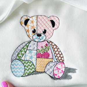 May include: Embroidered teddy bear with a patchwork design. The bear features various patterns, including stars, stripes, and floral motifs, in shades of pink, blue, green, and brown. The bear is sitting on a white surface.