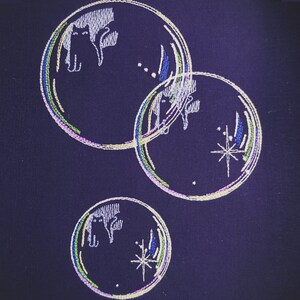 May include: Three embroidered circles on a dark purple fabric. Each circle has a white outline and a star in the centre. The circles are layered on top of each other, creating a sense of depth. The circles are decorated with colourful embroidery, including green, blue, and yellow threads.