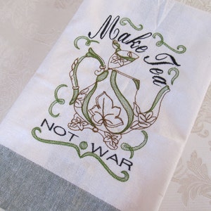 Pode incluir: Toalha de chá branca com um desenho bordado de bule verde e castanho. O texto "Make Tea Not War" é bordado em preto e verde. A parte inferior da toalha tem uma faixa cinzenta.