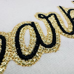 Puede incluir: Primer plano de un parche bordado con purpurina dorada con la palabra "party" en cursiva. Las letras están delineadas con hilo negro. El parche está sobre un fondo blanco.