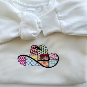 May include: White sweatshirt featuring an embroidered patchwork cowboy hat design in various colours.