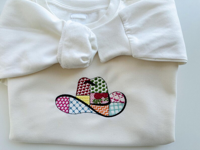 May include: White sweatshirt featuring an embroidered patchwork cowboy hat design in various colours.