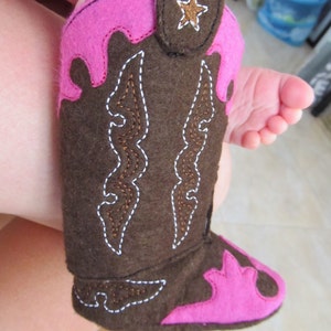 May include: A brown felt cowboy boot with pink felt accents and white stitching. The boot has a star on the side and a decorative design on the front.