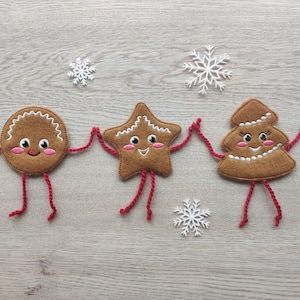 May include: Three felt gingerbread people holding hands. The gingerbread people are a round gingerbread man, a star-shaped gingerbread person, and a gingerbread tree. They are all smiling and have red yarn arms and legs. The gingerbread people are on a light brown wooden background.