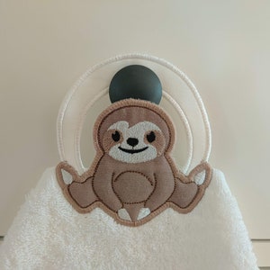 Puede incluir: Toalla de mano blanca con un aplique de perezoso marrón. El perezoso tiene una cara sonriente y está rodeado por un anillo blanco y una bola verde oscuro para colgar. La toalla está hecha de un material suave y absorbente.